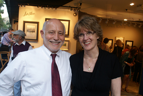 The image shows a man and a woman posing together at an art gallery, with both individuals smiling and looking directly at the camera.