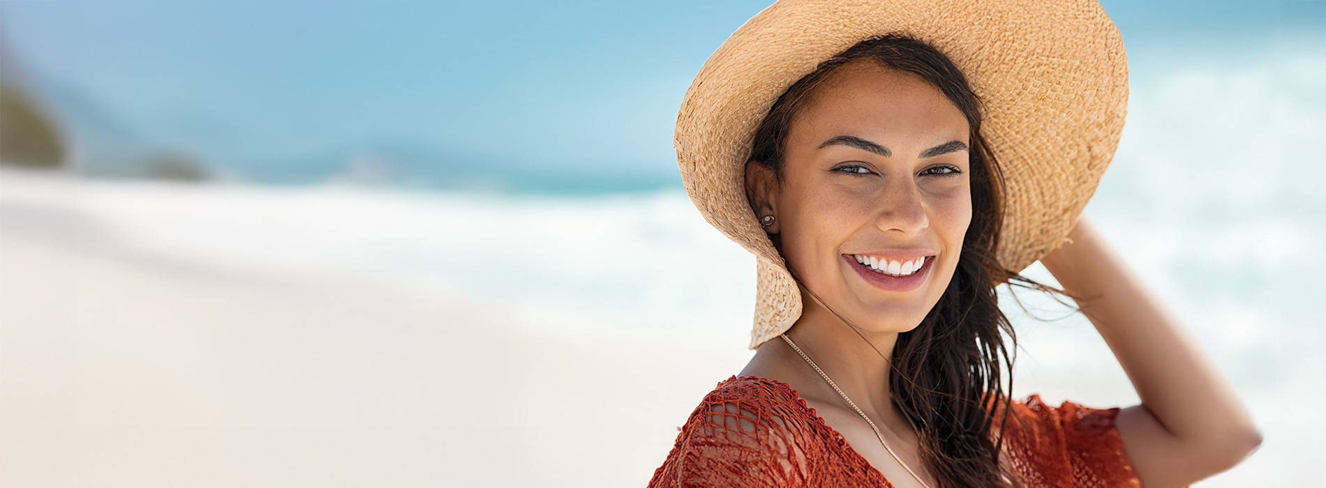 A smiling woman wearing a straw hat stands on a beach with a clear sky above her.