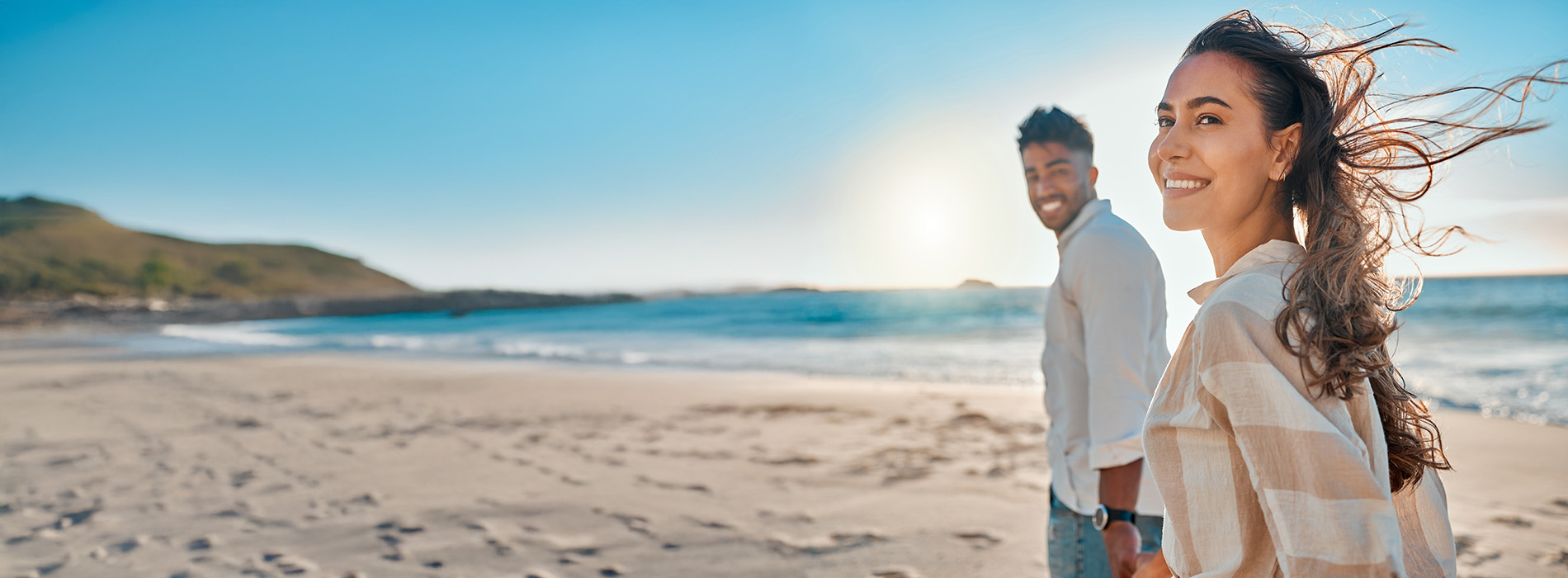 A photograph capturing two people walking along a sandy beach with clear skies above, under a bright sun, at sunset, with one person looking out towards the ocean.