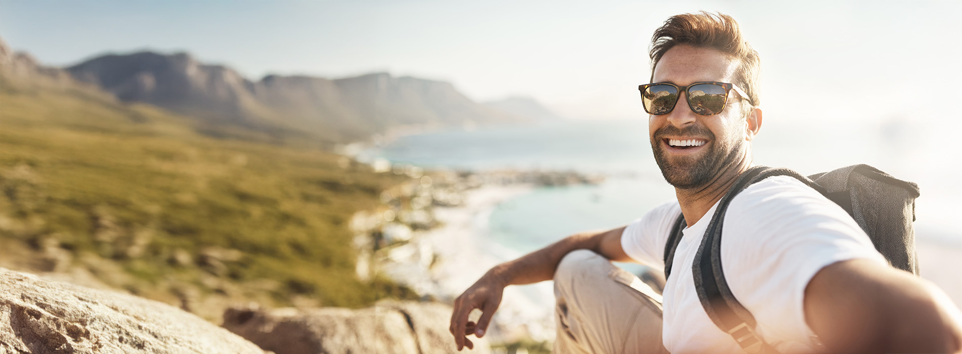 A man smiling at the camera while standing on a rocky outcrop with a mountainous landscape in the background, wearing sunglasses and a backpack.