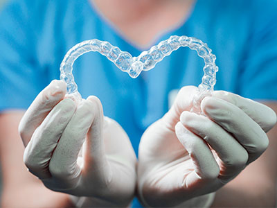 A person s hands holding a pair of clear dental braces with a heart shape inside, set against a blurred background featuring medical equipment.