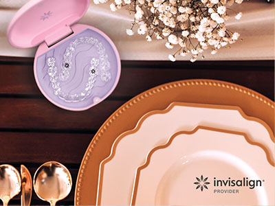 The image features a table setting with various dinnerware items including plates, bowls, and cutlery, along with a decorative box of matches and a piece of jewelry, all arranged on a wooden surface against a backdrop that includes a floral arrangement and a product packaging for  Invisalign.