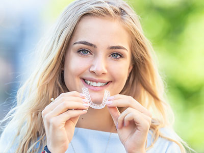 The image shows a smiling woman holding a toothbrush with toothpaste on her teeth, posing for a photo outdoors during daylight.