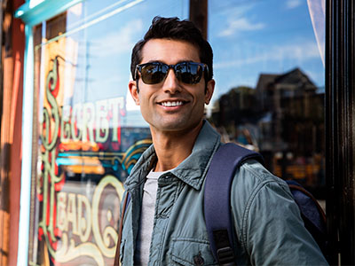 The image features a man standing in front of a store window, wearing sunglasses, a backpack, and a light-colored jacket. He has short hair, is smiling, and appears to be posing for the photograph.