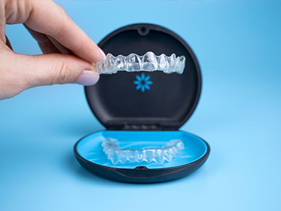 The image shows a person s hand holding an open black case containing a clear plastic dental aligner with blue braces, placed on a blue background.