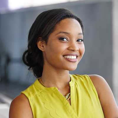 A smiling young woman with dark hair wearing a yellow top poses for a portrait against a blurred background.