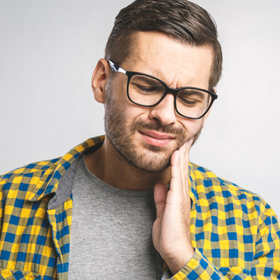 The image features a man with glasses and a beard, wearing a yellow plaid shirt, looking upwards with his hand on his chin, and he appears to be in pain or discomfort, possibly due to toothache or dental issues.