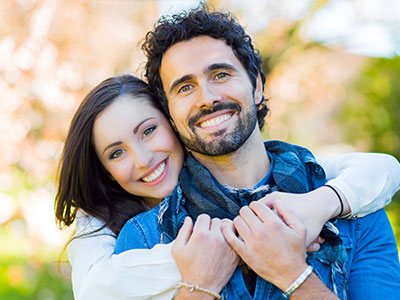 The image shows a man and a woman embracing each other with smiles on their faces  they are outdoors, under clear skies, and appear to be enjoying a warm, pleasant day together.