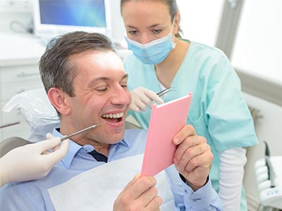 A man sitting in a dental chair with a cardboard sign over his face, receiving attention from a dentist who is smiling while holding a tablet.