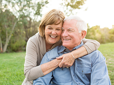 The image shows an older man and woman sharing a warm embrace outdoors during daylight.