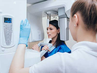 In the image, there are two individuals wearing blue gloves  one person is standing behind a large white machine with screens and buttons, while another person is seated at the machine, looking towards the camera. The setting appears to be an indoor environment, possibly a medical or scientific facility.