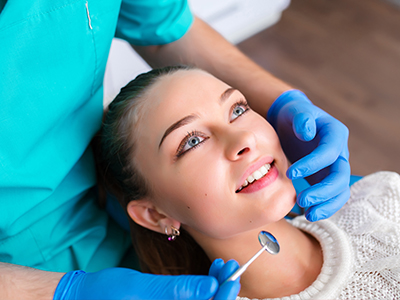 In the image, a woman with blue eyes is sitting in a dental chair, receiving dental care from a professional wearing a blue surgical mask and a white coat, who appears to be performing an examination or procedure on her teeth.