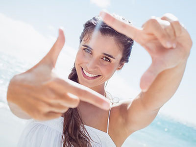 The image shows a smiling woman with long hair, wearing a white top, holding up her index finger against a bright blue sky background, with a clear focus on her action.